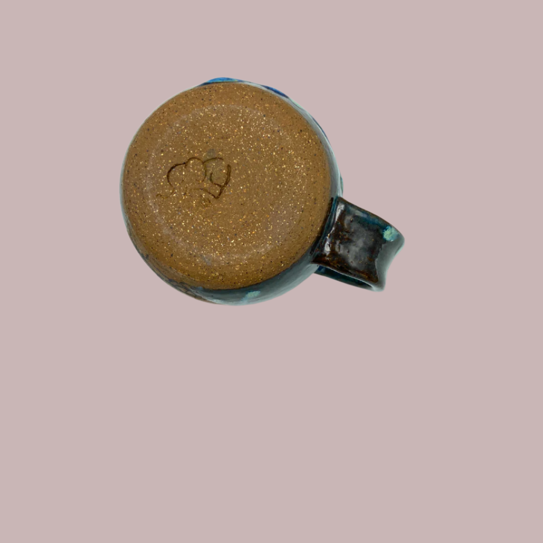 bottom view of A ceramic mug with a handcrafted tree design in blue and green colors on a brown clay background on cream colored background