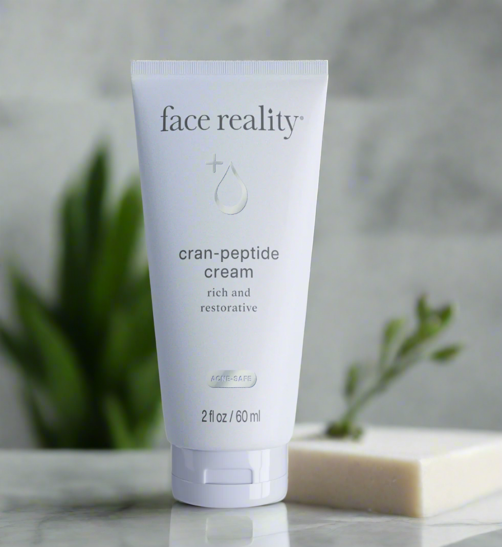 A tube of Face Reality Cran-Peptide Cream resting on a marble surface with a plant in the background.