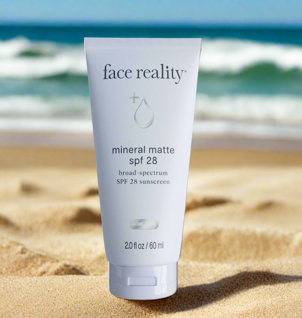 Tube of Face Reality Mineral Matte SPF28 sunscreen placed on sand with a beach and ocean in the background.