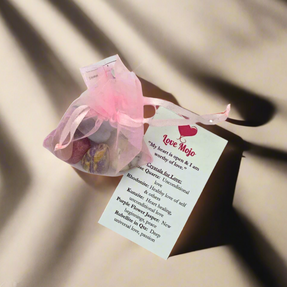 A pink organza bag tied with ribbon containing various small crystals, with a card labeled 'Love Mojo' explaining the product's purpose for manifesting love and enhancing emotional well-being.