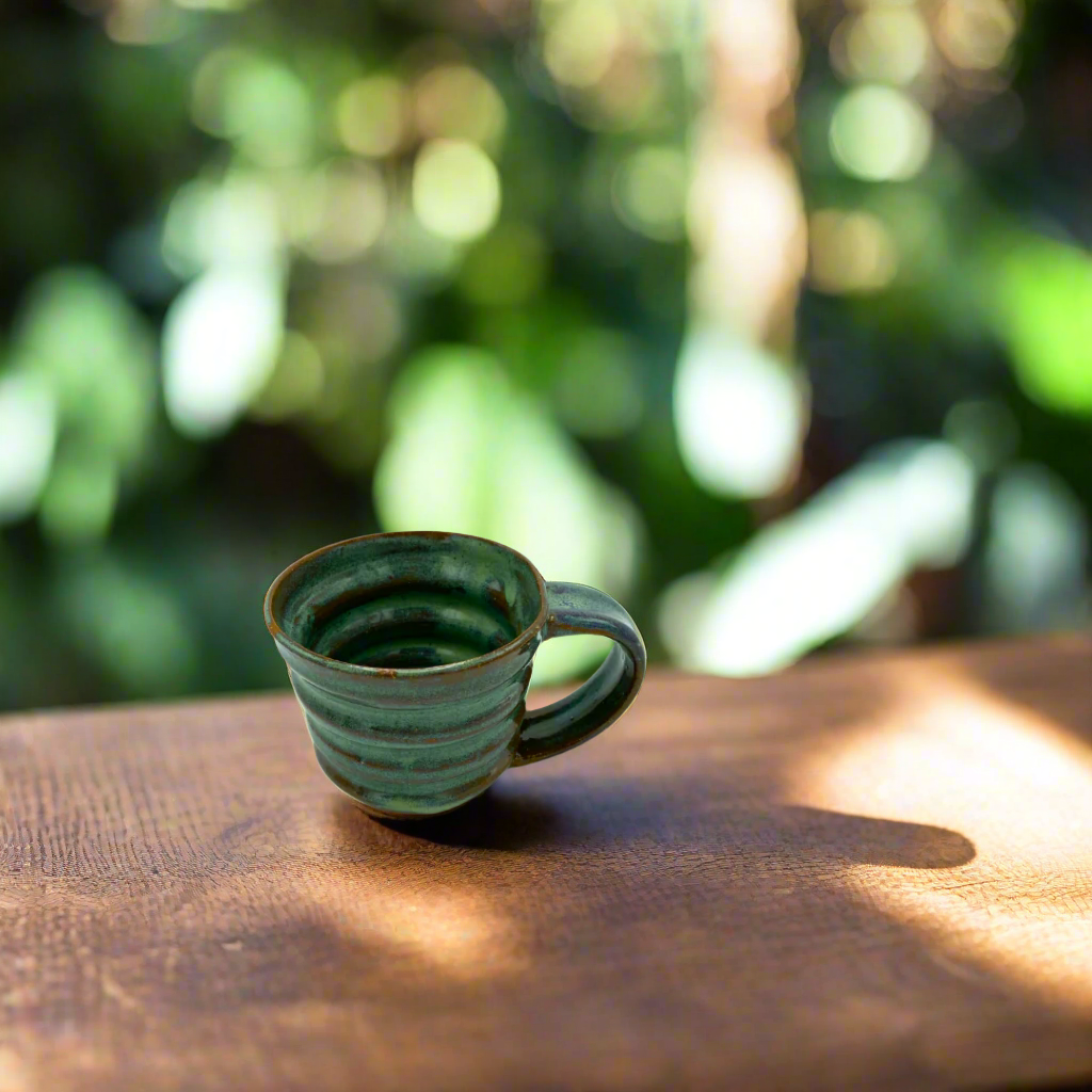 A handcrafted green ceramic mug with a textured surface and handle, placed on a wooden surface with a blurred greenery background.