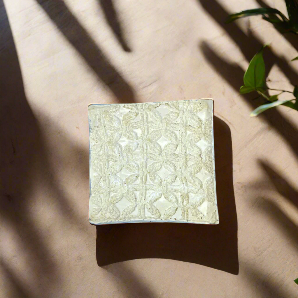 A beige square trinket dish with a floral pattern, placed on a surface with a shadow cast over it.