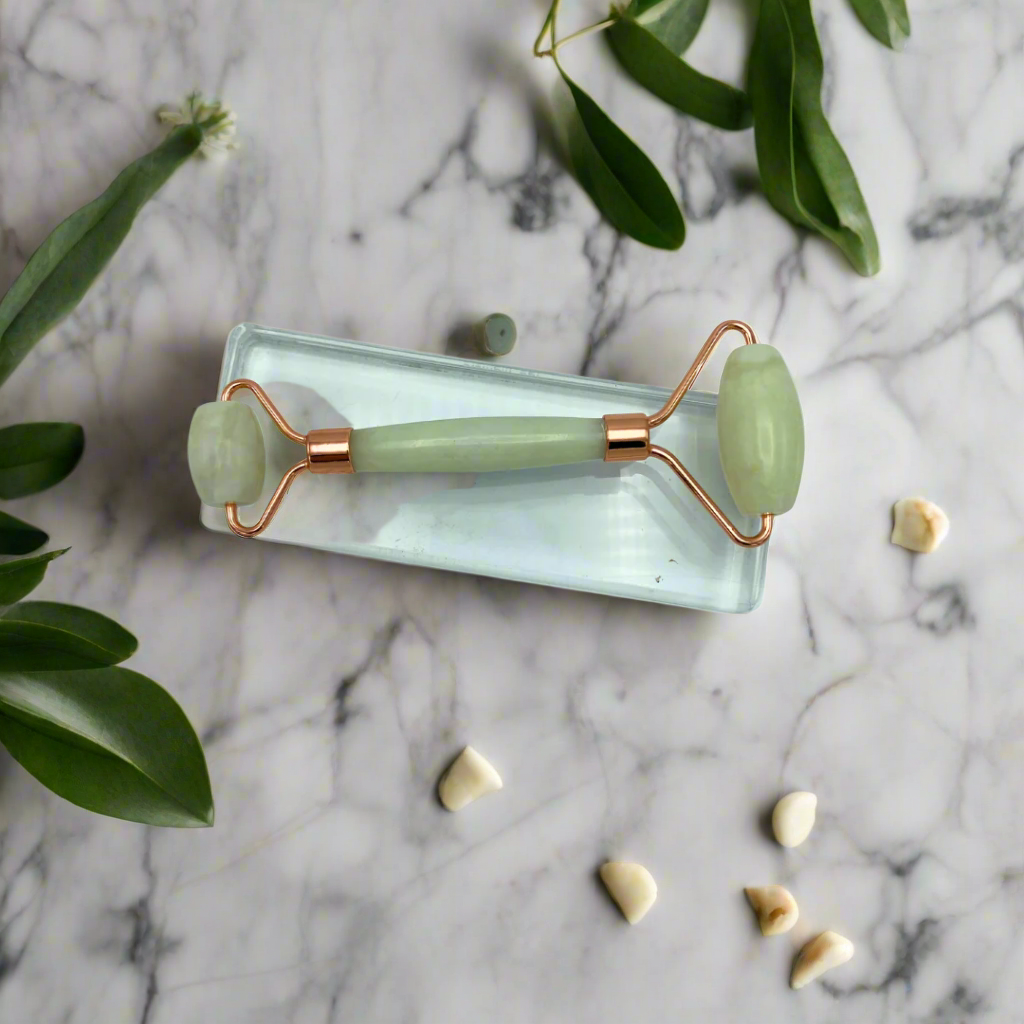A jade facial roller with copper-colored metal ends is displayed on a marble surface surrounded by leaves and small jade pieces.