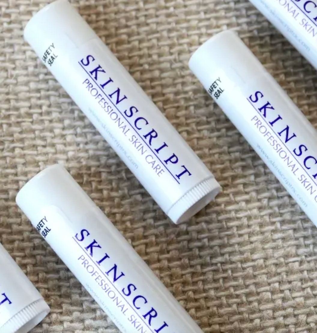 A collection of white lip balm sticks with the brand name 'SKINSRIPT' printed in blue on each stick, displayed on a textured background.